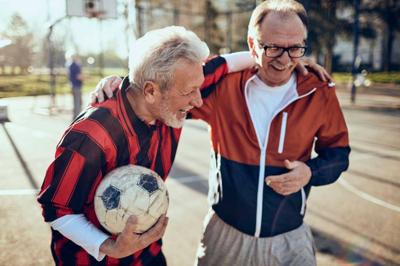 Zwei gute gelaunte Rentner in Sportkleidung, einer davon hat einen Fußball in der Hand.