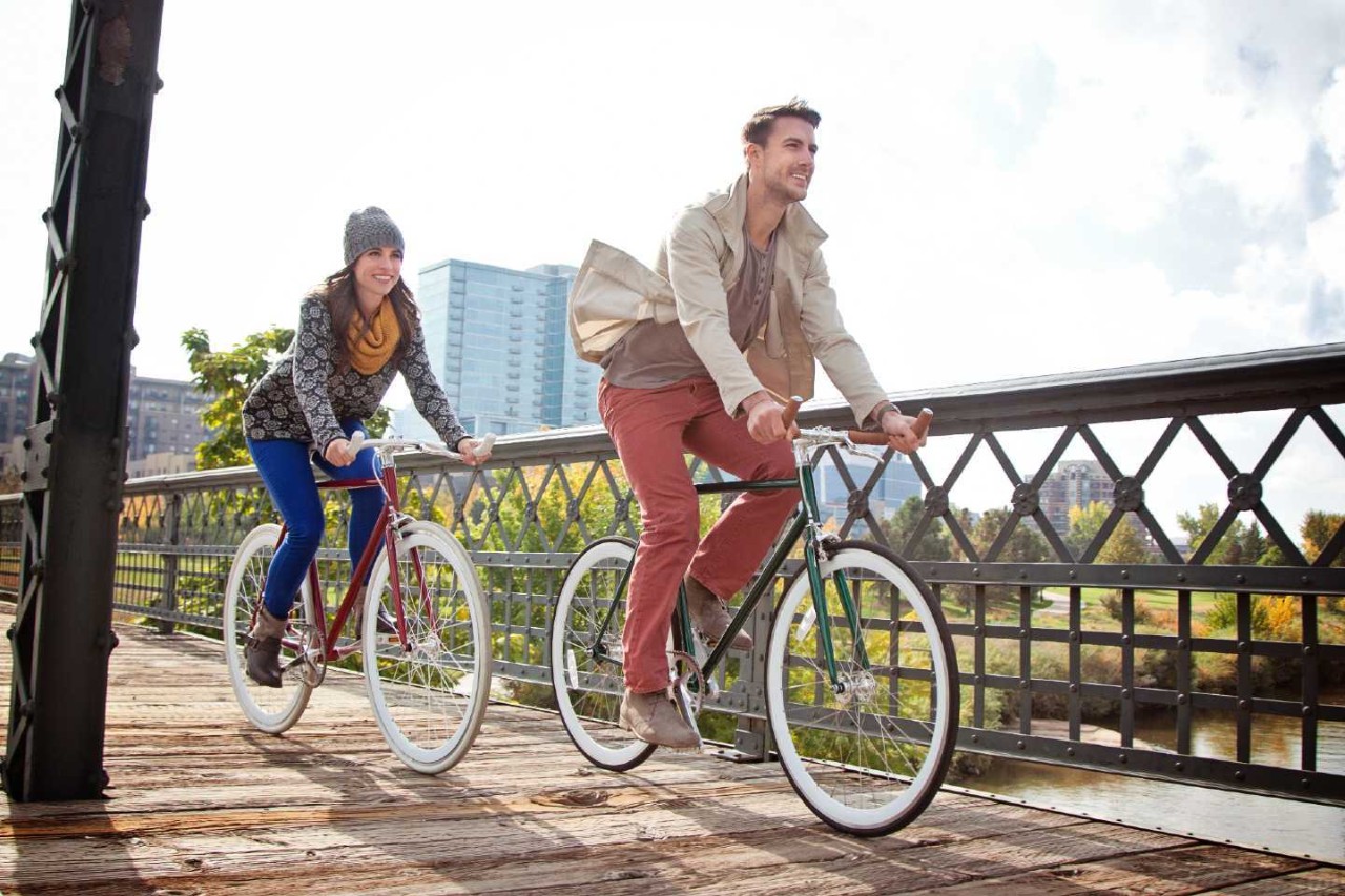Junge Frau und junger Mann fahren mit Fahrrädern über eine Brücke.