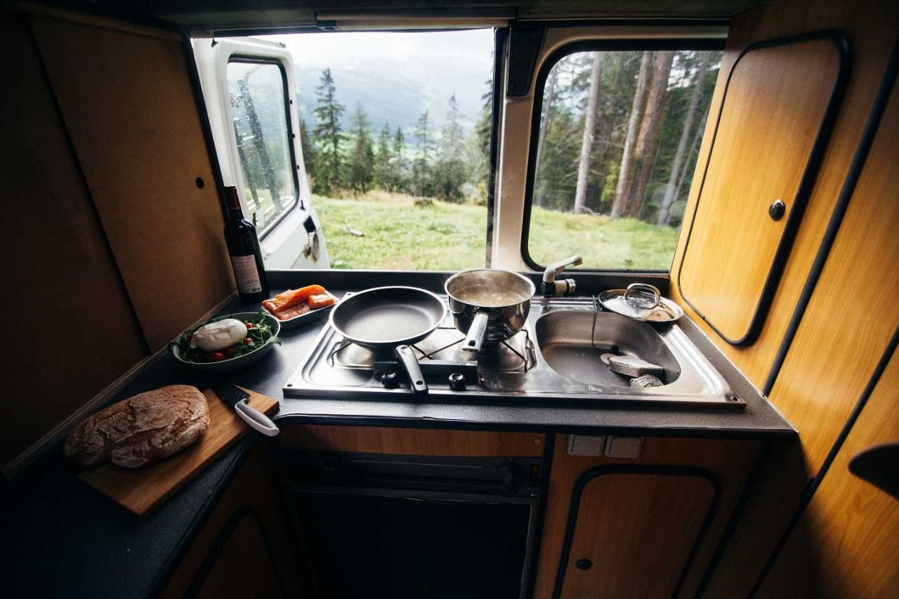 Auf dem Herd einer Camper-Küche steht eine Pfanne und ein Topf. Links daneben liegt ein Brot auf einem Holzbrett und ein Salat mit Mozzarella.