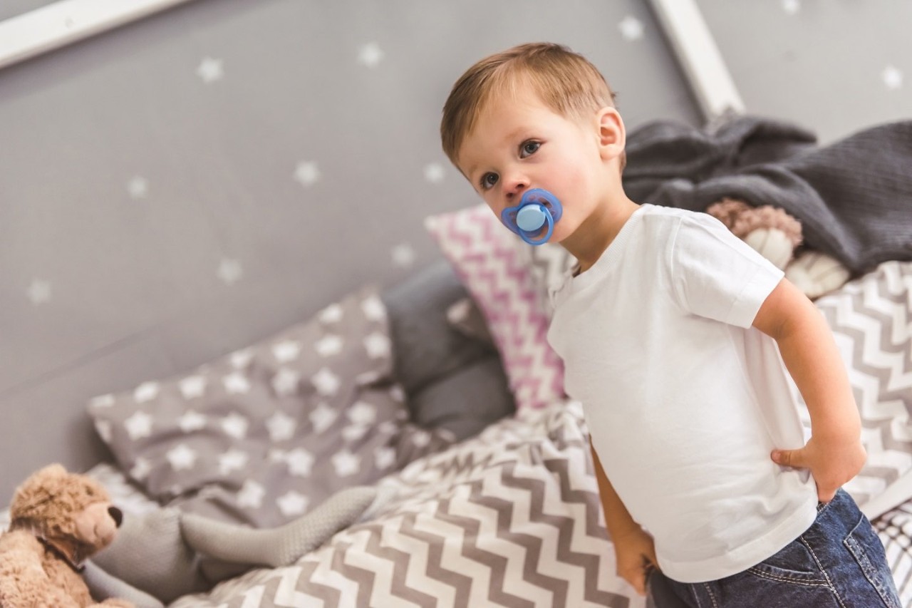 Ein Kleinkind mit Schnuller im Mund steht vor einem Bett.
