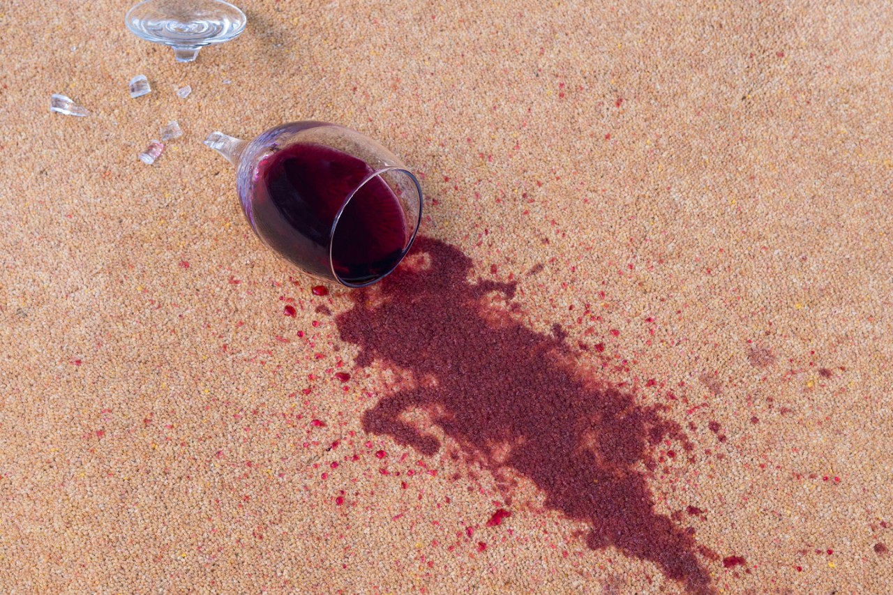 Ein zerbrochenes Weinglas neben einem Rotweinfleck auf dem Teppich.