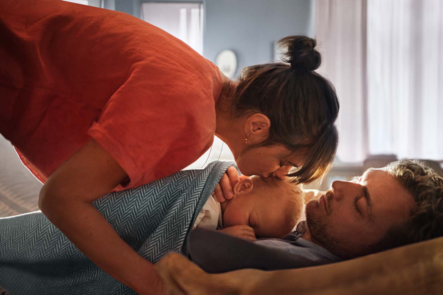 Ein Baby schläft auf der Brust des Vaters. Die Mutter gibt dem Baby einen Kuss auf den Kopf.