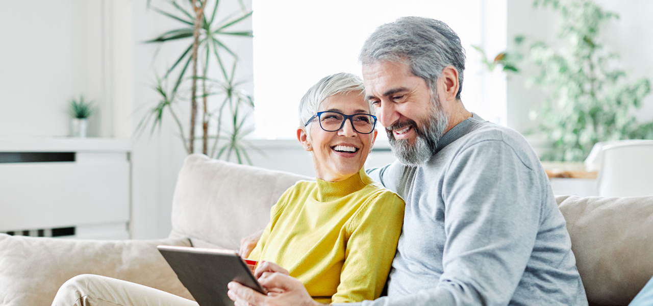 Älteres Paar sitzt lachend auf dem Sofa mit einem Tablet in der Hand.