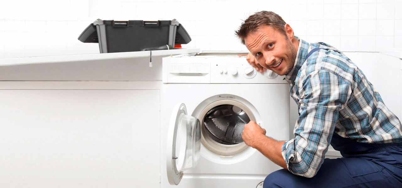 Ein Handwerker sitzt vor einer Waschmaschine und sieht direkt in die Kamera. 