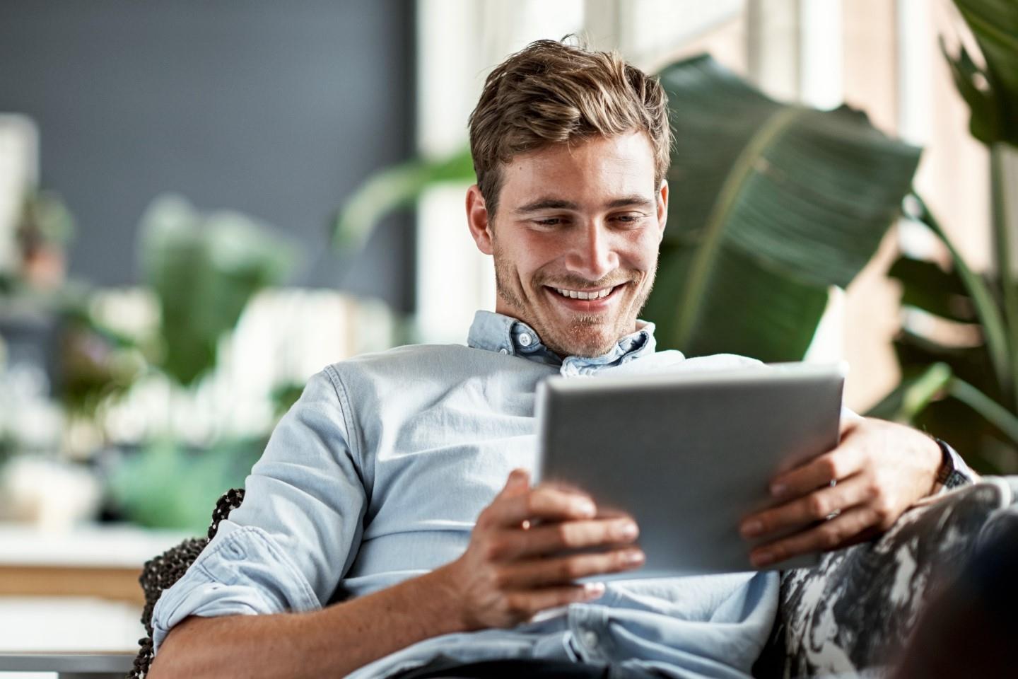 Ein junger Mann auf dem Sofa lächelt über das, was er im Tablet sieht.